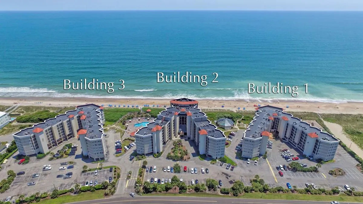 This exterior scene captures three distinct resort buildings facing a spacious swimming pool, with the sandy beachfront and ocean visible in the distance. The pool area is lively yet relaxing, hinting at community gatherings and family fun under the sun. Several balconies overlook the pool and ocean, reinforcing the connection between leisure and breathtaking views.