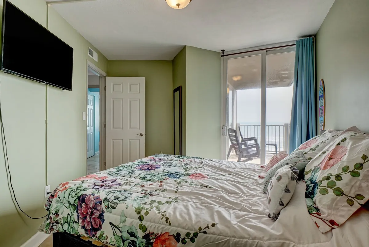 A tranquil bedroom with floral bedding, a large window overlooking the ocean, and a wall-mounted TV. The room feels peaceful and inviting, offering a serene retreat for reading or unwinding while enjoying the coastal scenery.