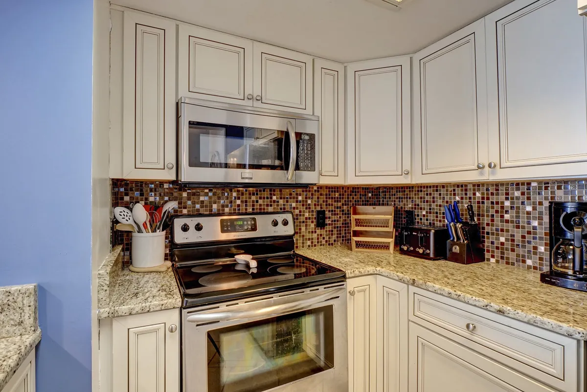 This image presents a modern kitchen with stainless steel appliances, a granite countertop, a mosaic backsplash, and a variety of kitchen utensils. The space appears bright and inviting, ideal for both cooking and entertaining guests. The open layout connects seamlessly to adjacent living or dining areas, encouraging interaction and togetherness.