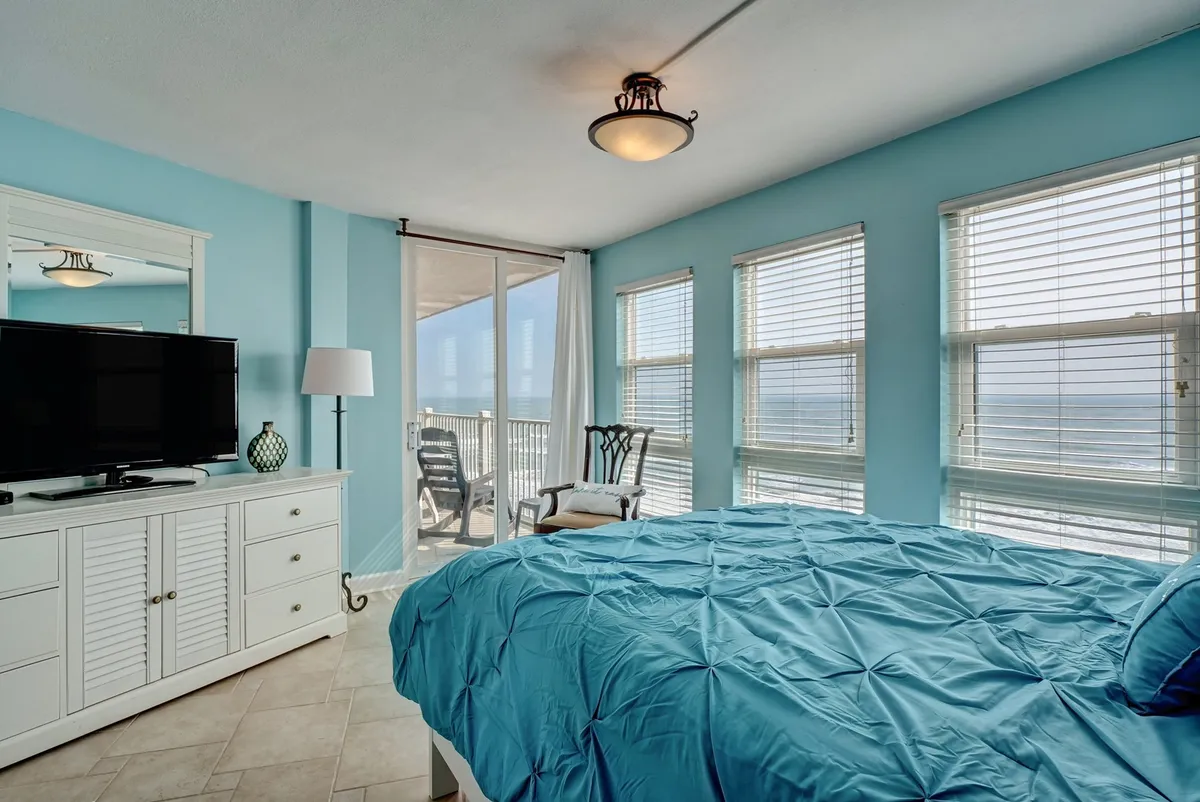 A spacious master bedroom featuring a large bed with blue bedding, a white dresser topped with a TV, and several windows that showcase an expansive ocean view. A cozy chair sits by the window, inviting relaxation and quiet moments.