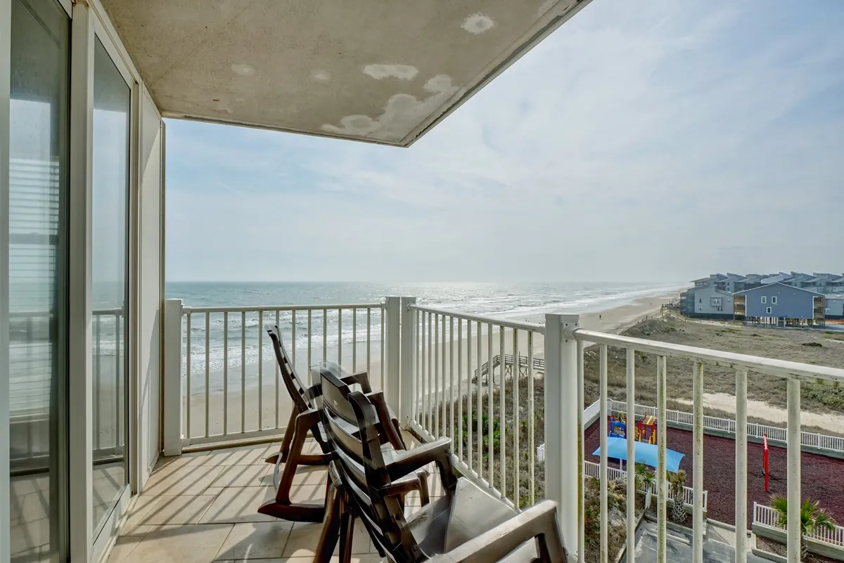 A private balcony with two chairs overlooking the expansive ocean and sandy beach. The inviting scene captures the tranquil blue of the sea meeting the sky, with gentle waves visible in the background. The balcony provides a direct connection to the outdoors, ideal for a serene work or relaxation spot.