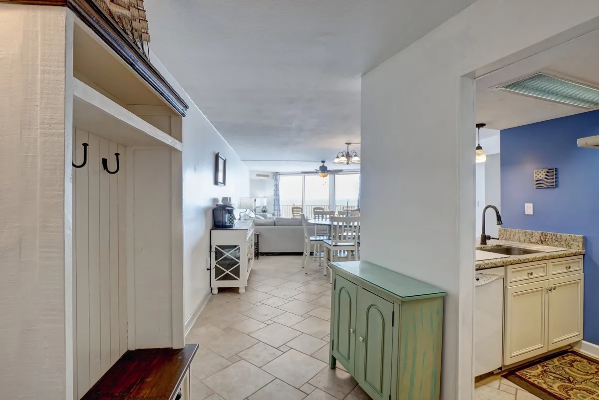 A view into the open-concept interior showing the entryway’s seating flowing seamlessly into the kitchen counter and dining area. Large windows fill the space with natural light and offer glimpses of the ocean, highlighting the layout’s welcoming and communal spirit.