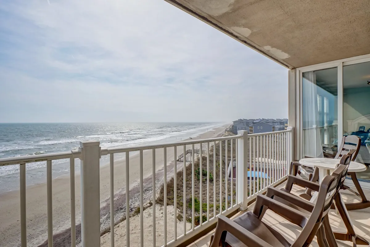 An exterior view from a private balcony with comfortable furniture, overlooking a tranquil ocean and sandy beach. The soft hues of sunrise cast a romantic glow across the water, setting a peaceful and intimate mood.