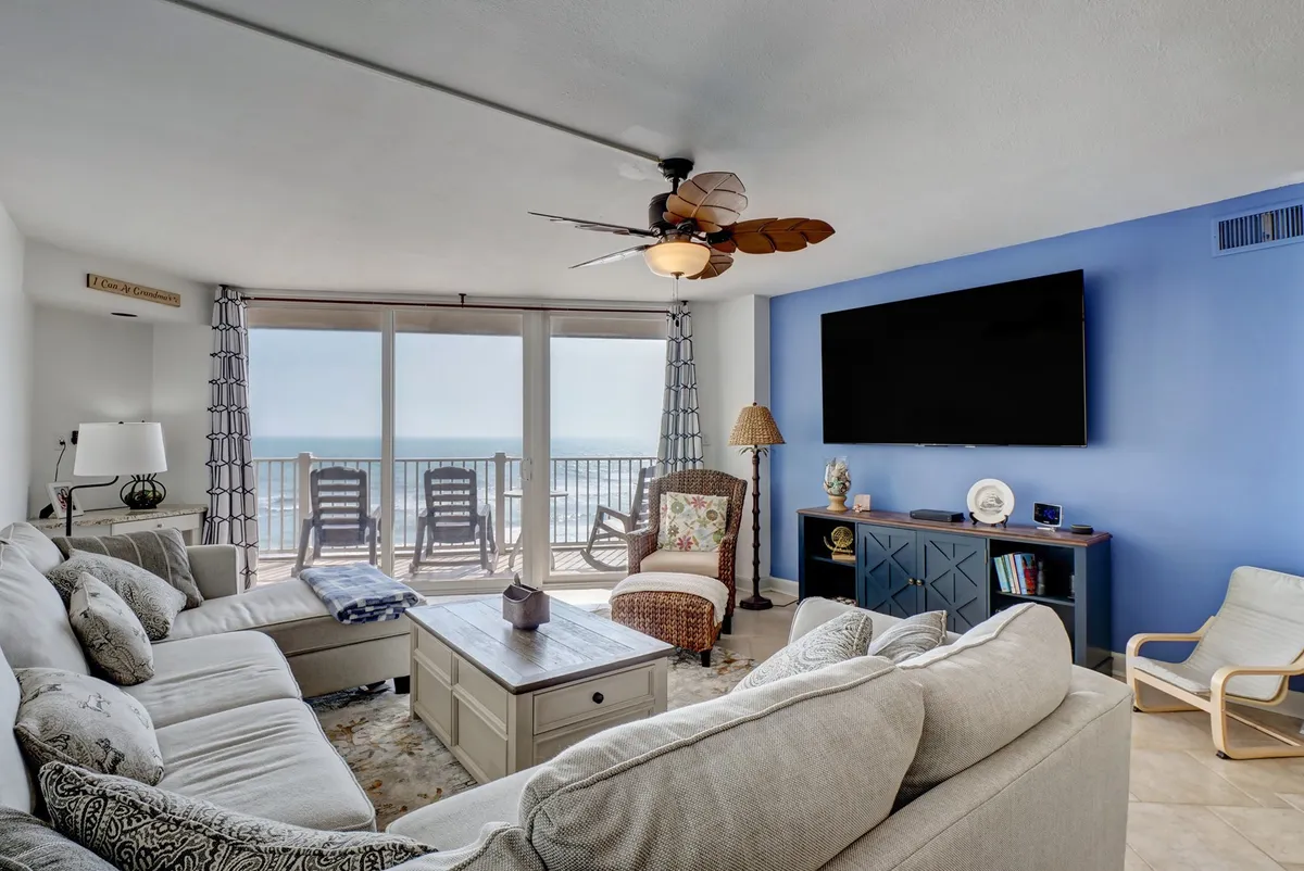 A spacious living room with a large sectional sofa positioned to face a wall-mounted TV. Floor-to-ceiling sliding glass doors offer a dramatic view of the ocean, seamlessly blending indoor comfort with the beauty of the Atlantic. Decorative lamp and coffee table elements create a cozy, inviting setting.