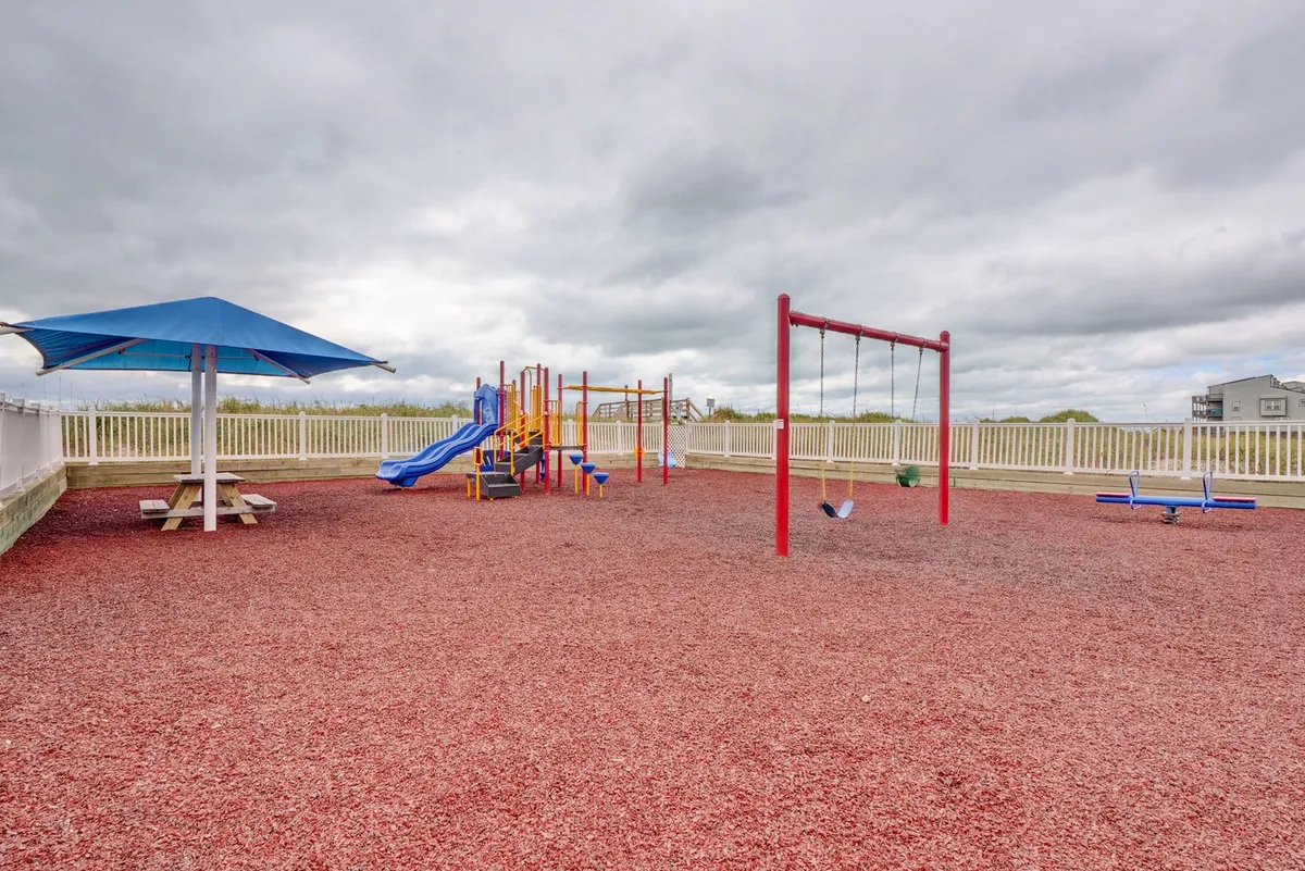A colorful playground equipped with swings, a slide, and a shaded picnic table sits on a green lawn, inviting children and families to play and relax. The casual, joyful atmosphere is perfect for family picnics, birthday parties, and leisure time.