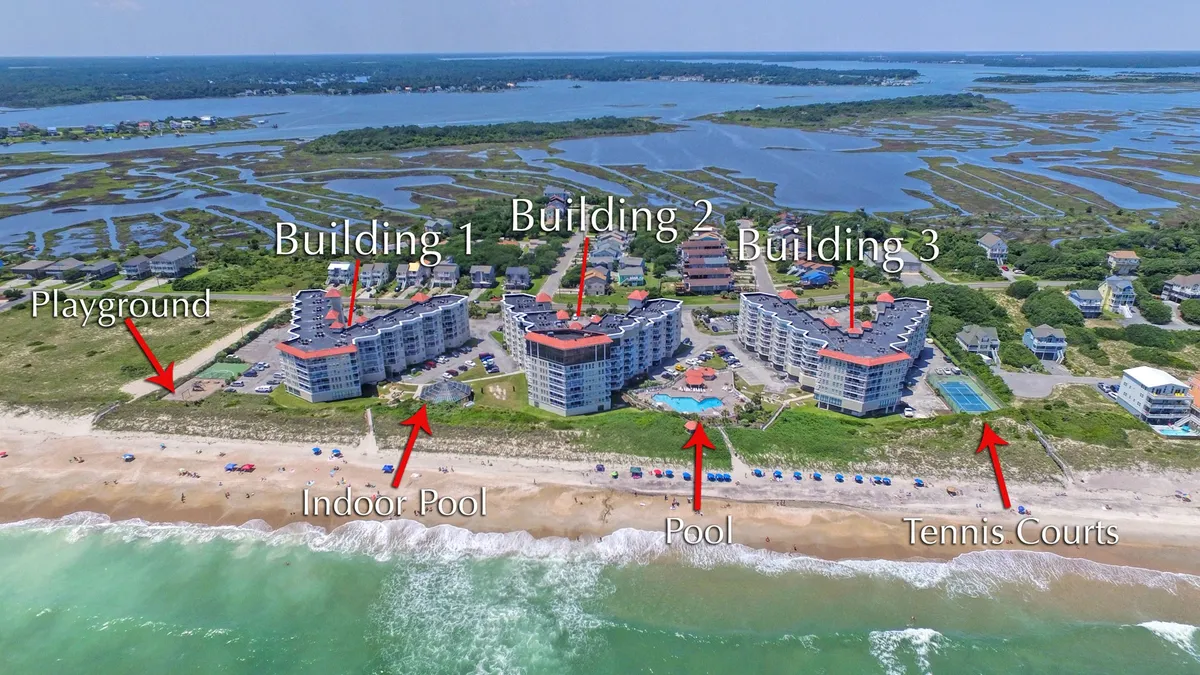 This exterior shot encompasses a wide view of the resort, showcasing three main buildings, the indoor and outdoor pools, tennis courts, playground, and beach access. The pools are focal points, while the sports courts and playground feature prominently, capturing the atmosphere of active recreation and relaxation.